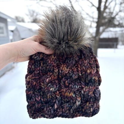 Knitted beanie with a fur pom-pom held in a hand against a snowy background